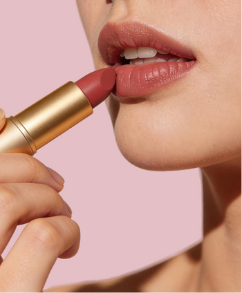 Close-up of a person applying a muted rosy nude lipstick from a gold tube, with the lips coated against a soft pink background.