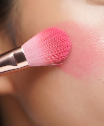 A close-up of a pink makeup brush applying bright pink blush to a person's cheek, showing the product being worn.