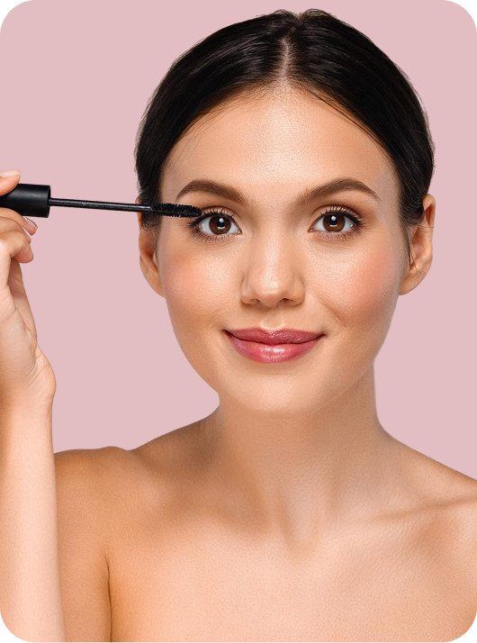 A close-up of a woman applying mascara to her upper lashes against a solid light pink background.