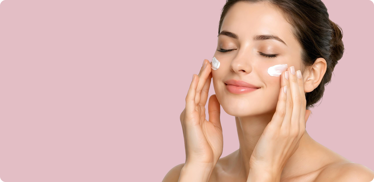 A woman with closed eyes applies white cream to her cheeks with her hands, set against a solid pink background.