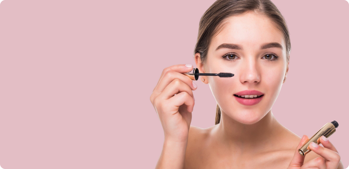 A woman with clear skin applies mascara to her upper lashes while holding a gold and black mascara tube in her other hand, set against a solid light pink background.